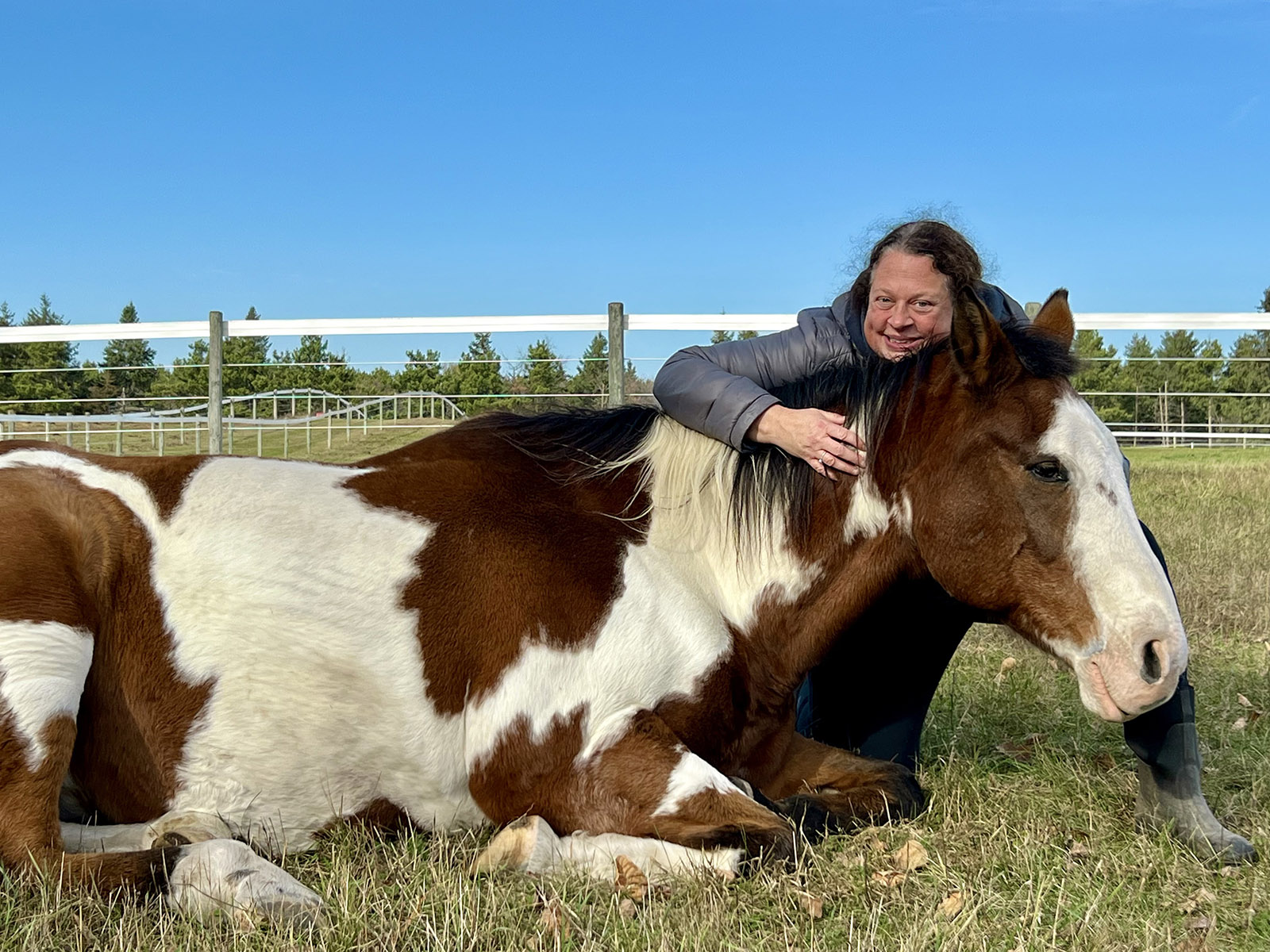 Smiling woman and horse sitting in a field with her arm around the horse